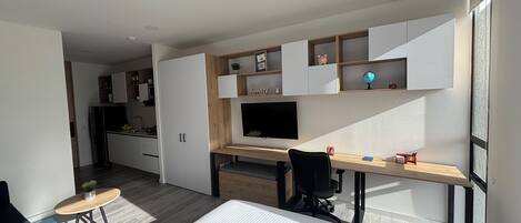 Un dormitorio moderno con cama, escritorio y televisor. Se observa una cocina al fondo.
