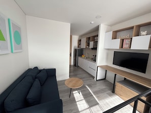 Uma sala de estar moderna com um sofá de cor escura, uma mesa de madeira redonda e uma TV fixada em uma prateleira de madeira.