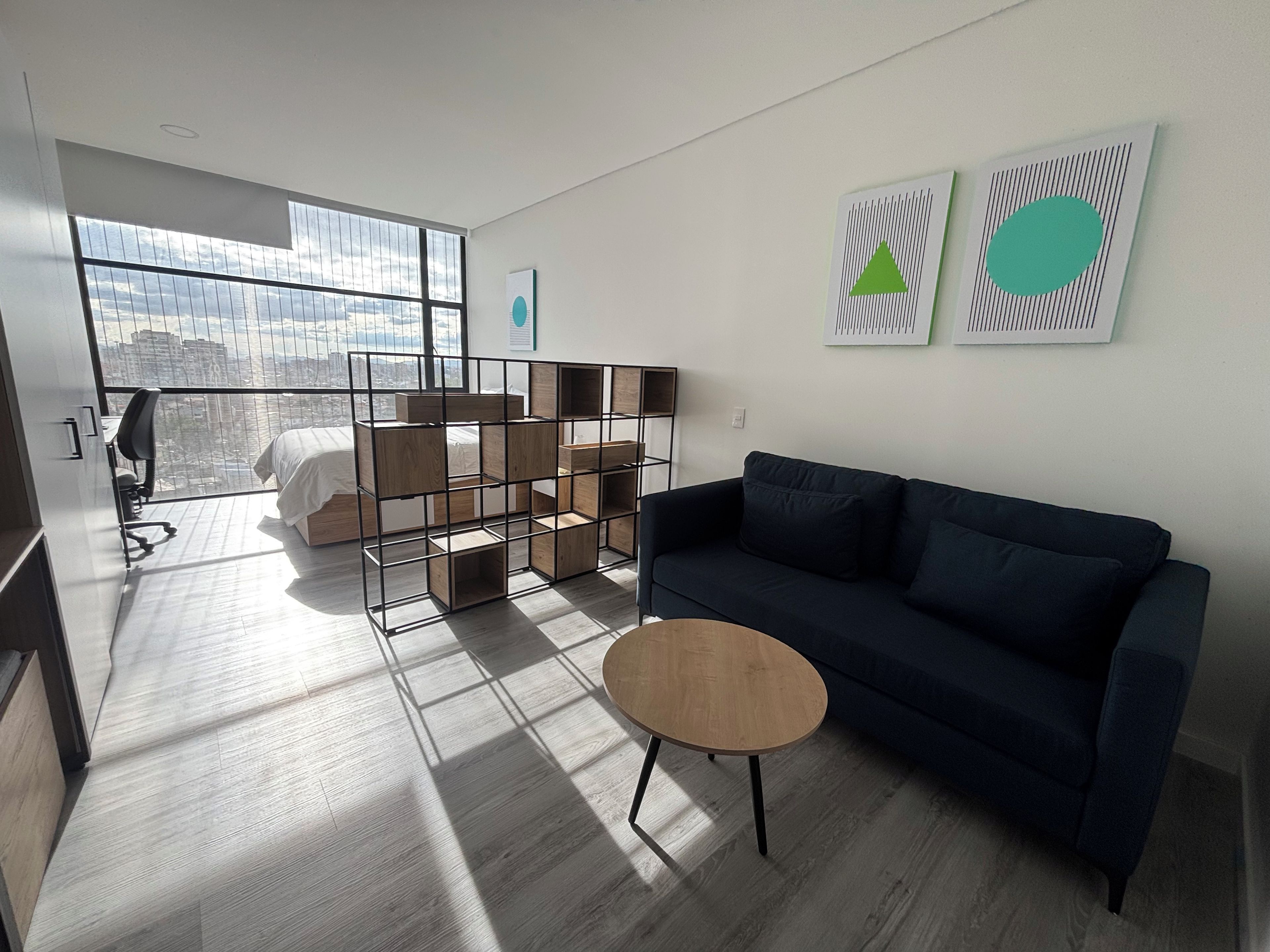 A modern hotel room with a large window, a dark blue sofa, a wooden coffee table, and a metal shelving unit.