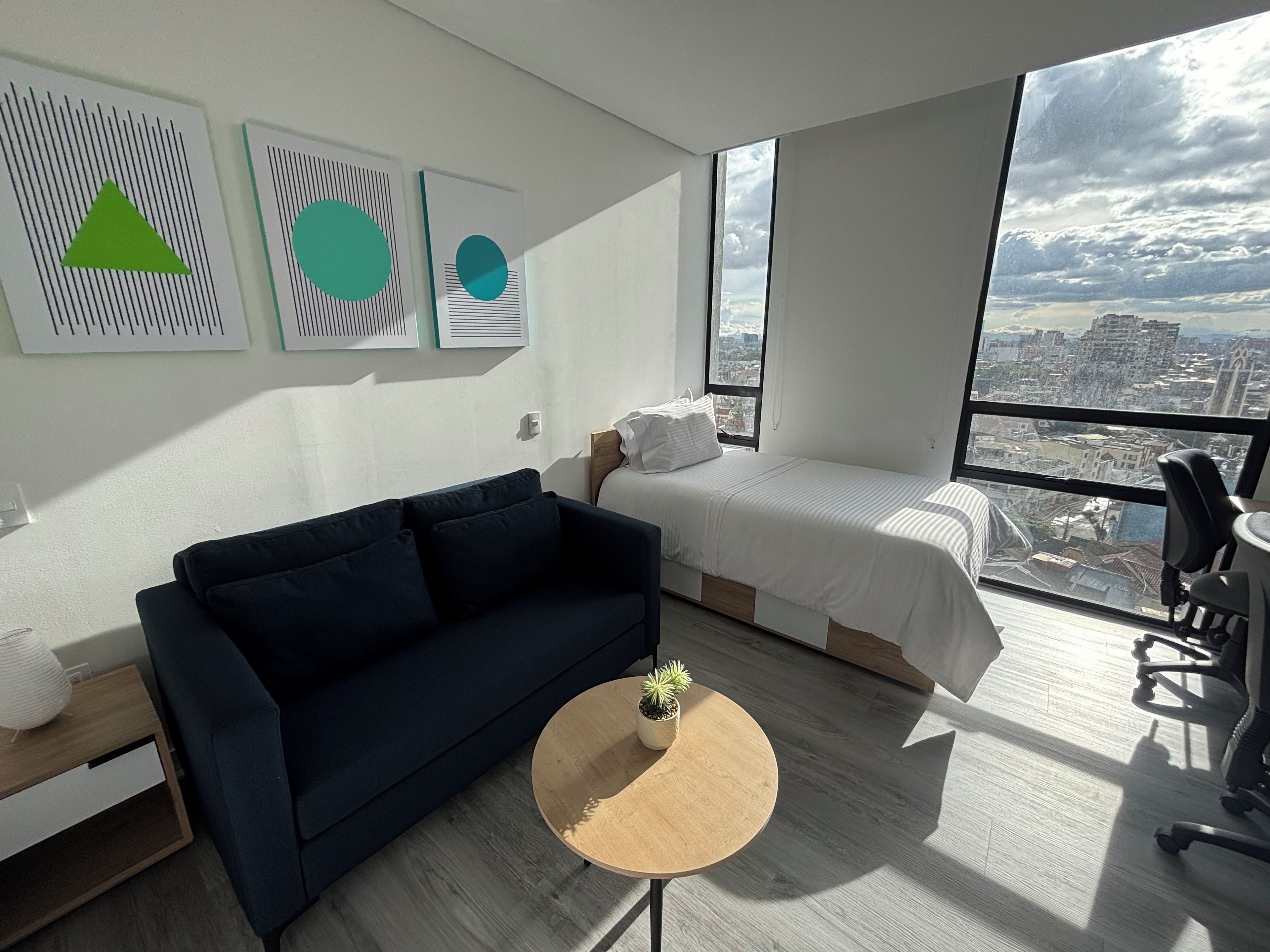 Une chambre d’hôtel moderne avec un grand lit, un canapé, une petite table ronde agrémentée d’une plante, et une vue sur la ville par une grande fenêtre.