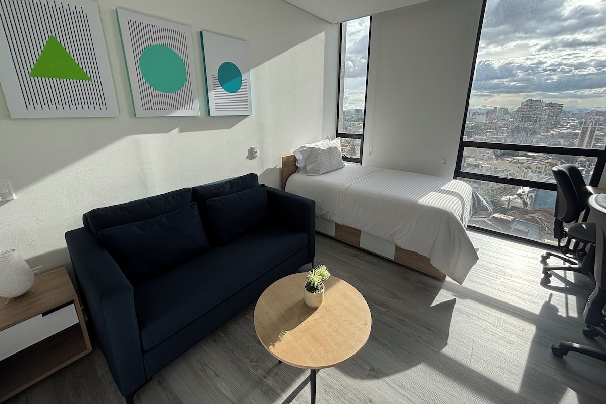 A modern hotel room with a large bed, a sofa, a small round table, and a view of the city through a large window.
