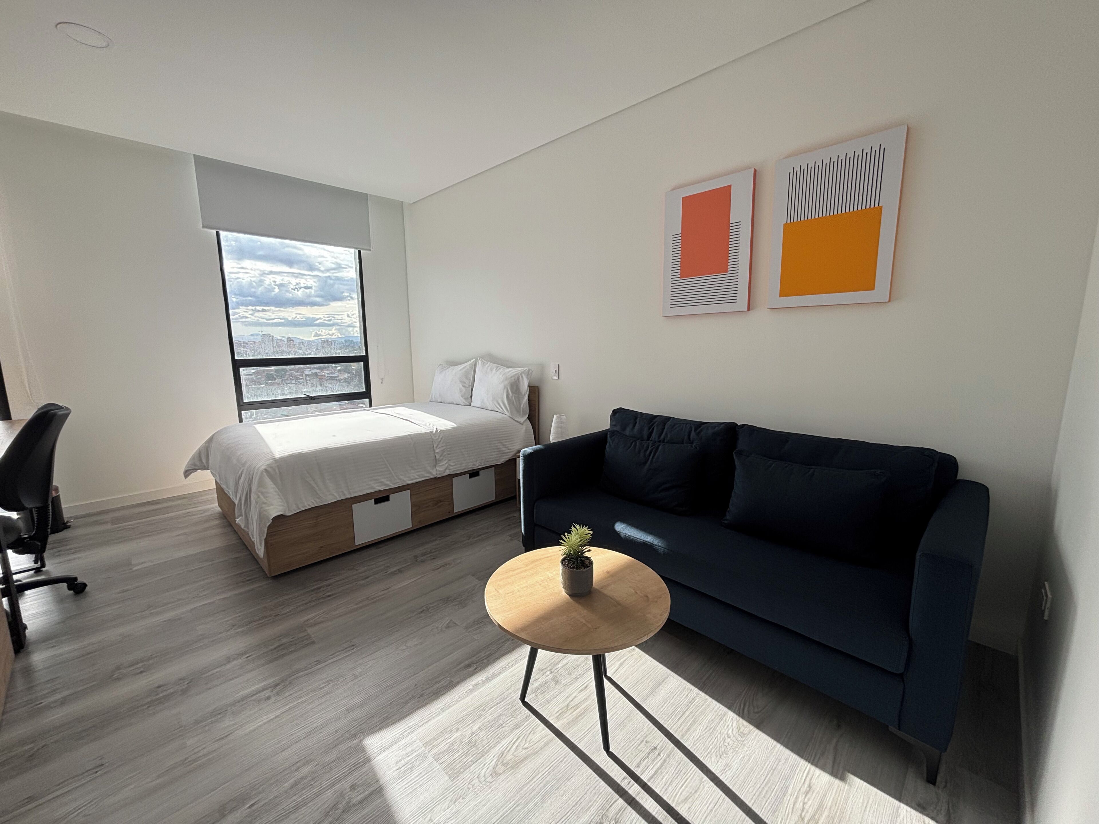 A modern bedroom with a bed, a dark blue sofa, a small round table, and a large window offering a city view.