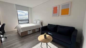 A modern bedroom with a bed, a dark blue sofa, a small round table, and a large window offering a city view.
