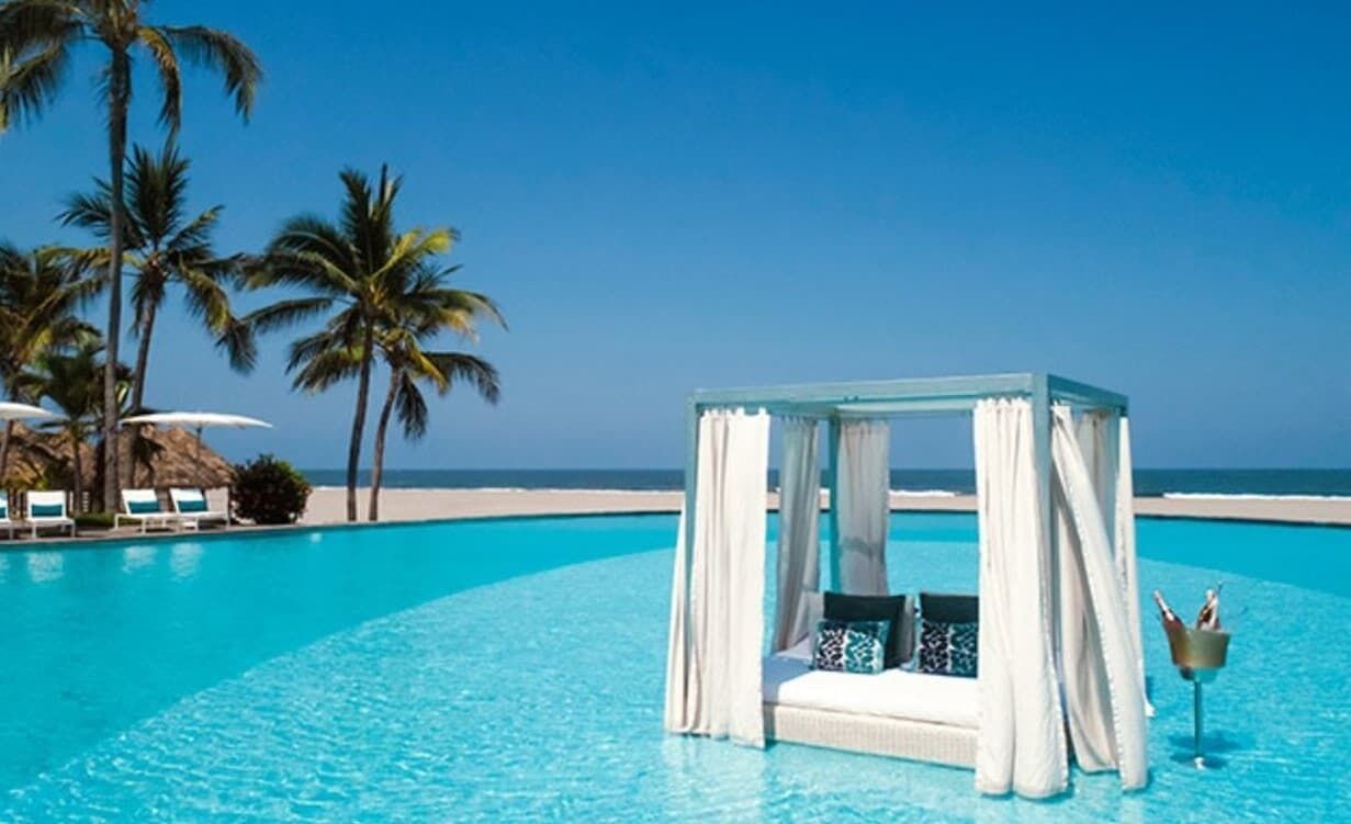 A clear blue pool with a white canopy gazebo, palm trees, and a beach in the background.