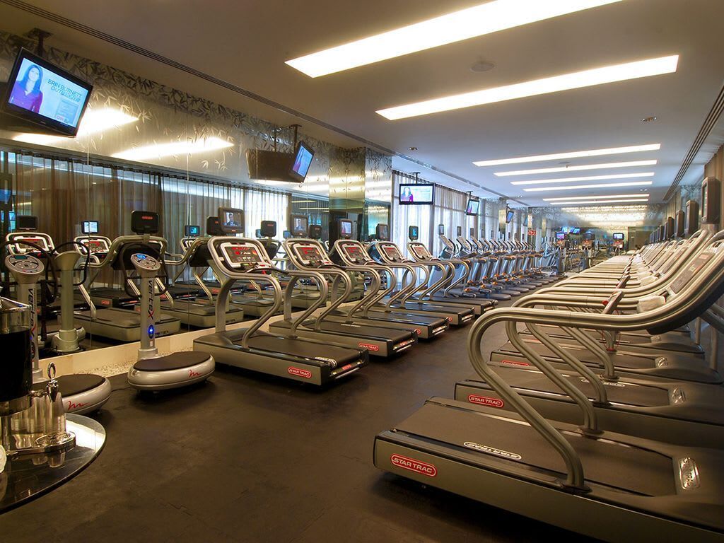 Ein Fitnessraum mit mehreren in Reihen angeordneten Laufbändern, einem an der Wand befestigten Fernseher und einem Spiegel, der den Raum reflektiert.