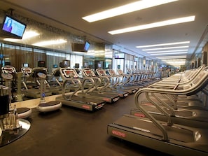 Ein Fitnessraum mit mehreren in Reihen angeordneten Laufbändern, einem an der Wand befestigten Fernseher und einem Spiegel, der den Raum reflektiert.