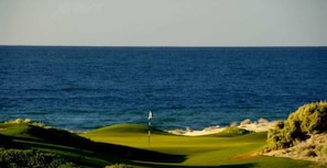Un parcours de golf avec un mât de drapeau sur le green, entouré de bunkers de sable et de végétation, avec l’océan en arrière-plan.
