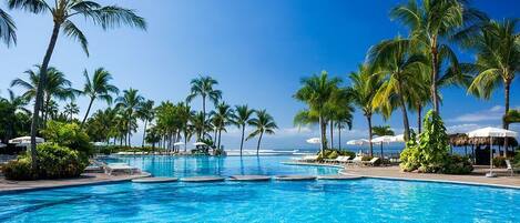 A large outdoor swimming pool with clear blue water, surrounded by palm trees and lounge chairs.