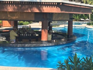 Un espace bar situé au bord de la piscine, avec un barman et un client, le tout entouré d’une piscine et de chaises longues.