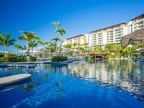 Ein Hotelpool mit klarem, blauem Wasser, umgeben von Palmen und einem mehrstöckigen Hotelgebäude.