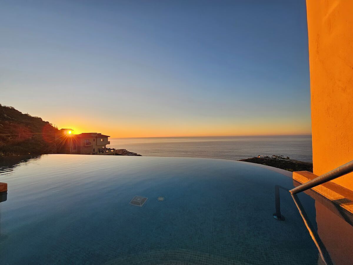An infinity pool with a view of the sunset over the sea, a building on the left, and a railing on the right.