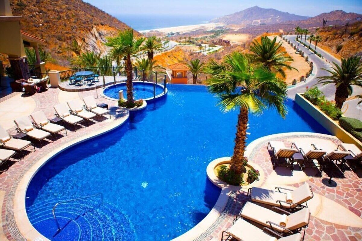 A resort pool with sun loungers and palm trees, surrounded by a mountainous landscape.