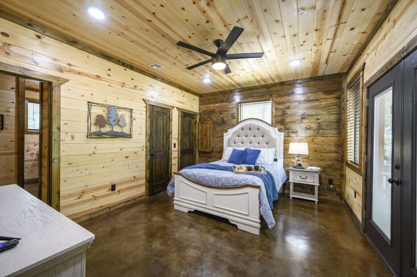 Une chambre avec un plafond en bois, un lit doté d’une tête de lit capitonnée, une table de chevet et une fenêtre avec des stores.