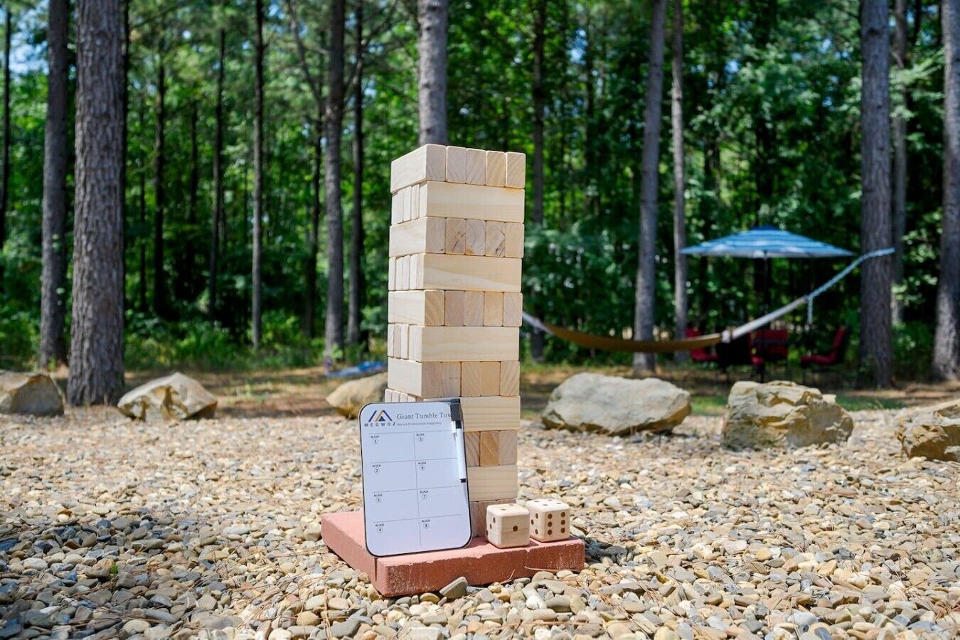 Un jeu de construction consistant à empiler des blocs de bois, avec une carte des règles et des dés posés sur un sol recouvert de petits cailloux, le tout entouré d’arbres.