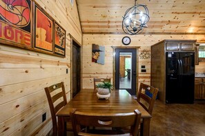 A wooden interior with a dining table, chairs, a clock, and a refrigerator.