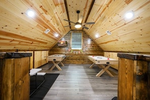 A wooden room with two foosball tables, a ceiling fan, and a window with blinds.
