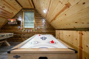 A wooden-floored room with a shuffleboard table, a foosball table, and a window with blinds.