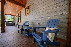 A wooden deck with Adirondack chairs and a table, a covered porch area, and a view of greenery outside.