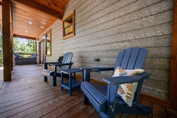 A wooden deck with Adirondack chairs and a table, a covered porch area, and a view of greenery outside.