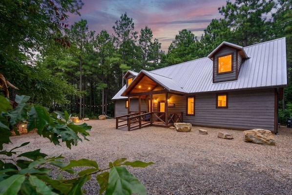A cabin with a porch, surrounded by trees and a gravel driveway.
