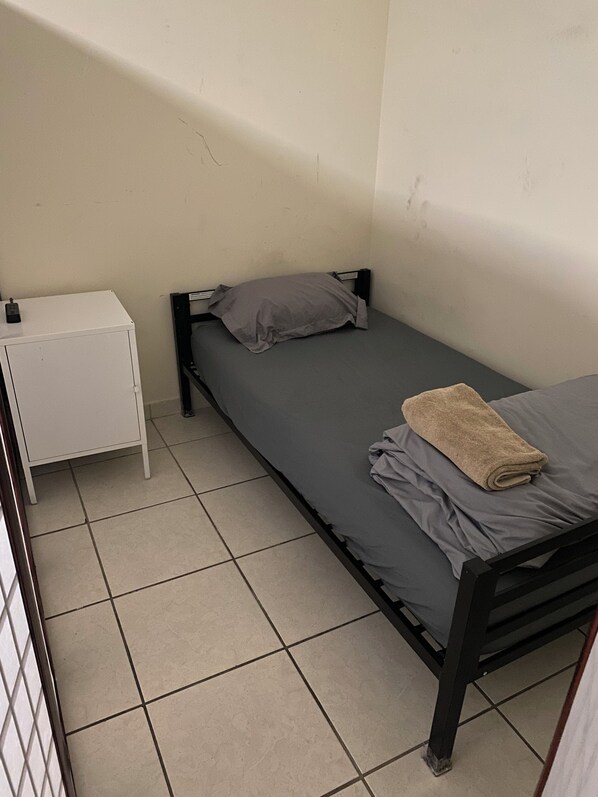 A small, single-bed room with a grey mattress and pillows, a white bedside table, and a brown towel on the bed.