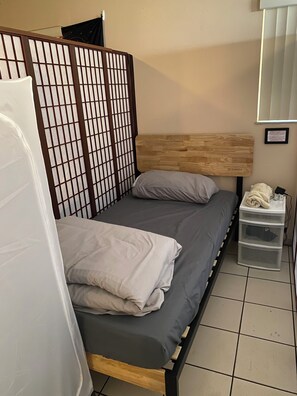 Una cama individual con cabecera de madera, ropa de cama gris y una cortina blanca parcialmente visible.