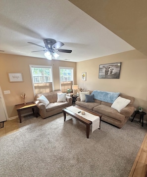A living room with a ceiling fan, two windows, a sofa, a coffee table, and a painting on the wall.
