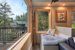 A cozy room with a wooden deck, a window with a view of trees, a couch with a pillow, and a framed picture on the wall. - Stylish 3BR Girdwood Home w Chugach Mountain Views (Anchorage)