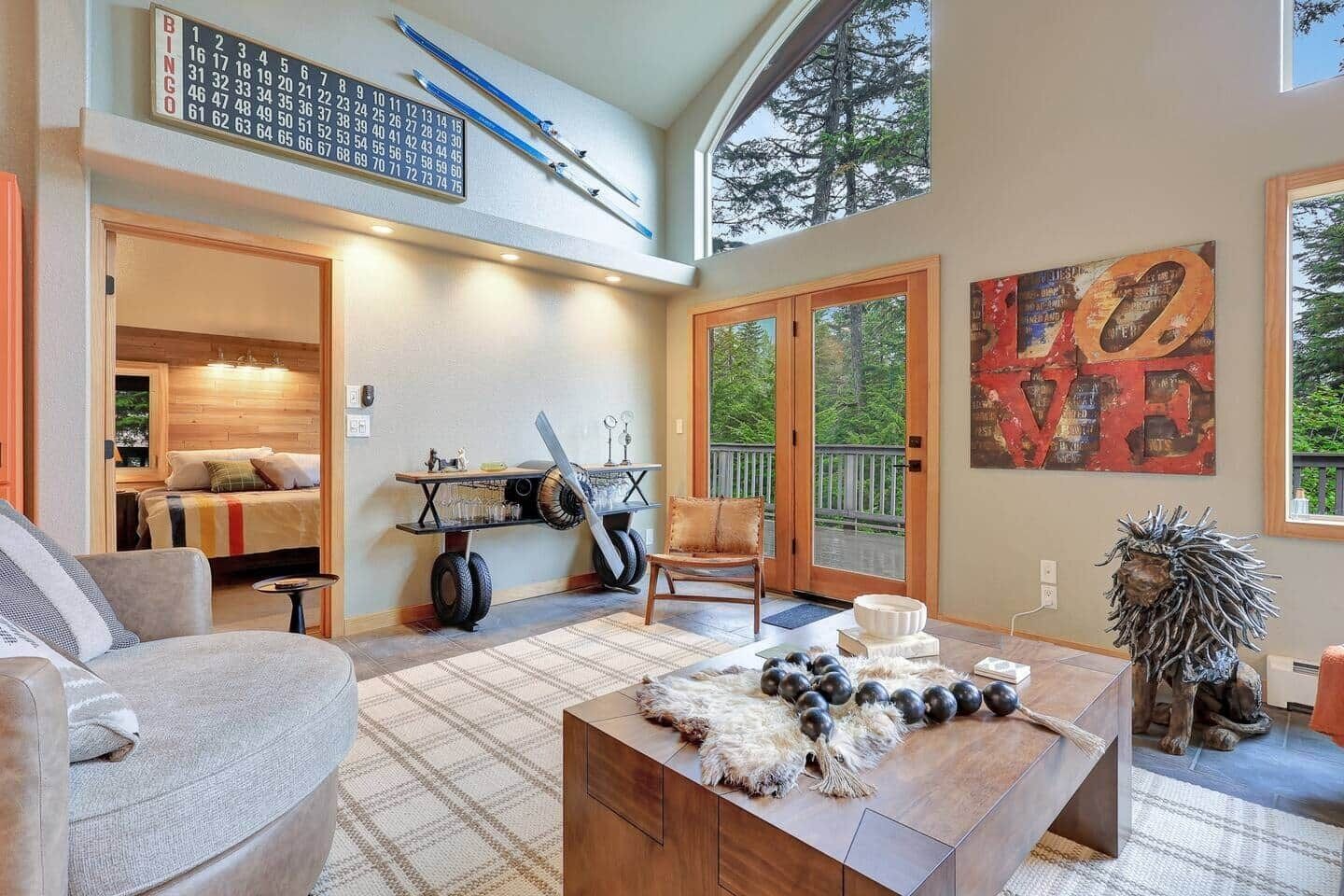 Amplia sala de estar con techos altos, un ventanal y vistas al exterior. Se observa una mesa de centro de madera con objetos decorativos, un sofá y una silla.