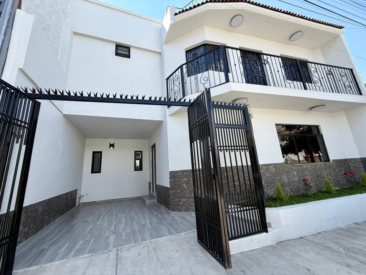 Un moderno edificio residencial de dos pisos con entrada protegida por un portón, un balcón en el nivel superior y un pequeño jardín con flores.