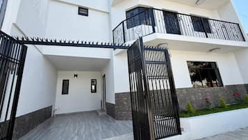 Un moderno edificio residencial de dos pisos con entrada protegida por un portón, un balcón en el nivel superior y un pequeño jardín con flores.
