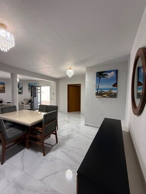 A spacious living area with a dining table, chairs, a black bench, a chandelier, and a painting of a beach scene.