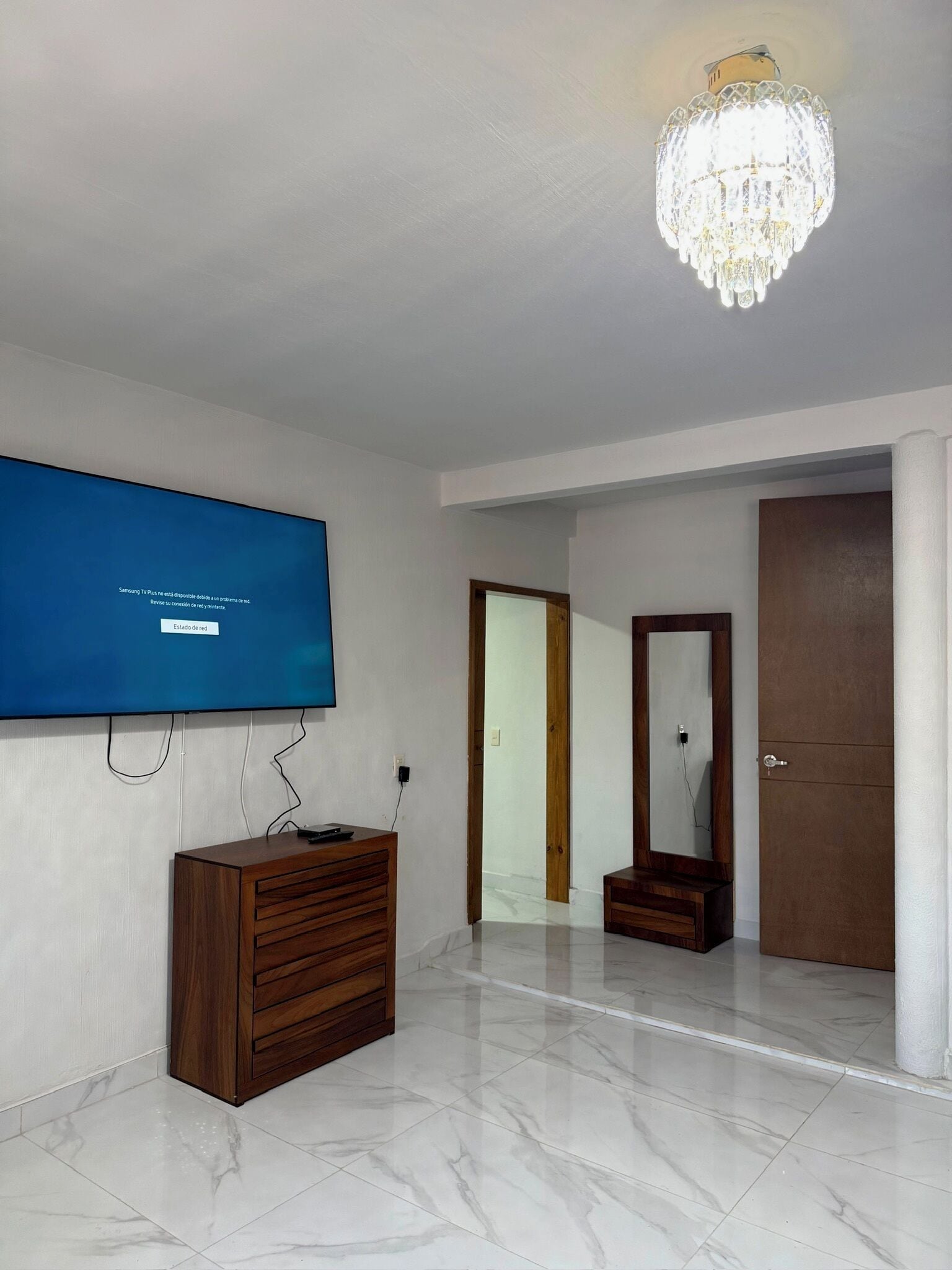 Una habitación con una televisión de pantalla plana mostrando un mensaje de error de red en rojo, un aparador de madera, piso de baldosas de mármol, una puerta de madera y un candelabro.