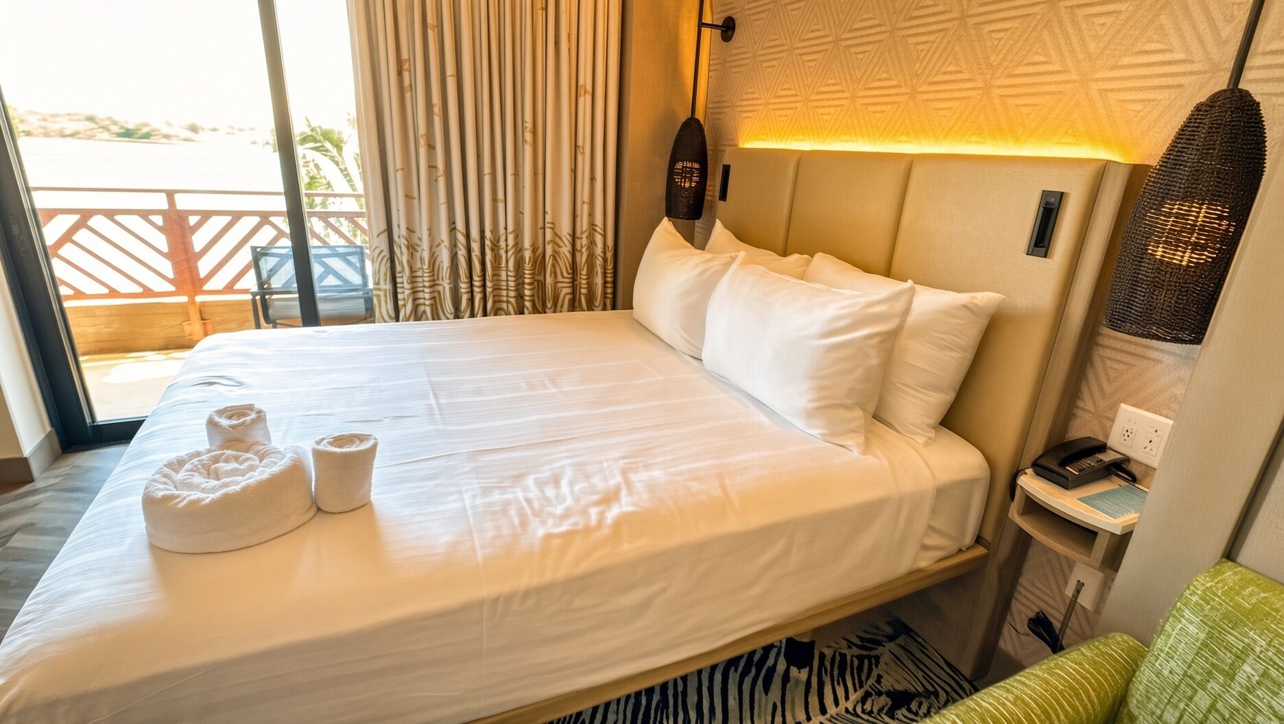 A hotel room with a bed, pillows, a bedside table with a lamp, a telephone, and a view of the outdoors.