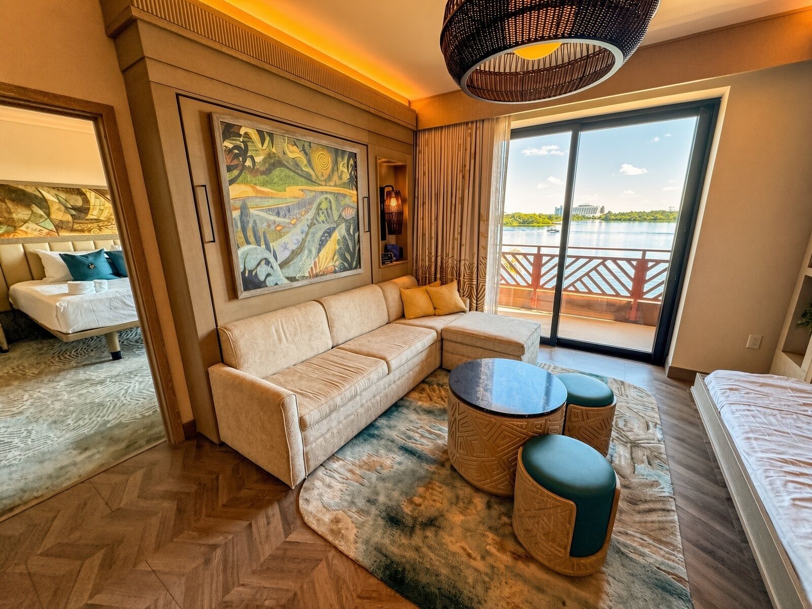 A hotel room with a sofa, a dining table, a large painting, and a view of a body of water through a sliding door.