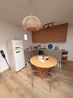 A modern kitchen with a white refrigerator, wooden cabinets, a round dining table with chairs, and a hanging light fixture. - Ferdinand's Cabin • Beach: 2 min • Private patio • Air Conditioning • Fiber (Arcachon)