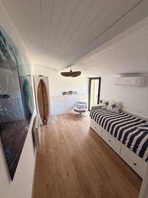 A bedroom with a bed, a chair, a wooden surfboard, and a wall-mounted air conditioner. - Ferdinand's Cabin • Beach: 2 min • Private patio • Air Conditioning • Fiber (Arcachon)