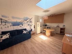 A modern kitchen with wooden cabinets, a dining area with a round table and chairs, a blue sofa with patterned cushions, and a beach-themed wall mural. - Ferdinand's Cabin • Beach: 2 min • Private patio • Air Conditioning • Fiber (Arcachon)