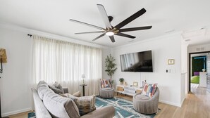 Un salón con sofá, sillones, mesa de centro, televisor, ventilador de techo y un amplio ventanal con cortinas.