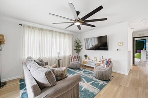 A living room with a sofa, armchairs, a coffee table, a TV, a ceiling fan, and a large window with curtains. - Anastasia Island Retreat - Villa B - Walk Downtown (St. Augustine)
