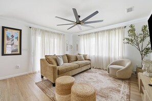 Ein Wohnzimmer mit einem beigefarbenen Sofa, einem runden Sessel, einem Deckenventilator und einem großen Fenster mit weißen Vorhängen.