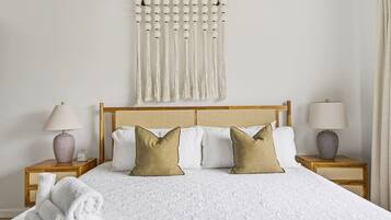 A neatly made bed with a white bedspread and two pillows, flanked by two bedside tables with lamps and a wall-mounted macrame curtain.