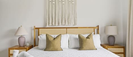 A neatly made bed with a white bedspread and two pillows, flanked by two bedside tables with lamps and a wall-mounted macrame curtain.