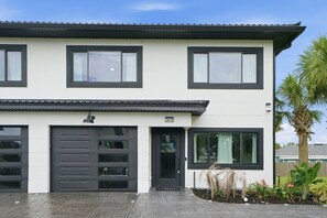 A modern two-story building with a garage, large windows, and a small garden with plants and a palm tree.