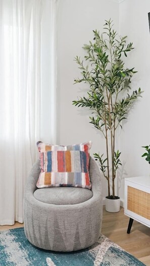 A room with a grey armchair, a colorful pillow, a tall potted plant, and a white cabinet. - Anastasia Island Retreat - Villa B - Walk Downtown (St. Augustine)