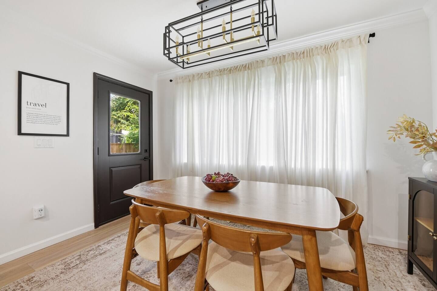 A dining area with a wooden table and chairs, a chandelier, a door, and a window with sheer curtains.