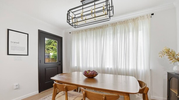 A dining area with a wooden table and chairs, a chandelier, a door, and a window with sheer curtains.