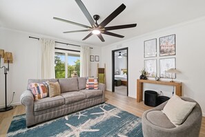Ein modernes Wohnzimmer mit Deckenventilator, einer Couch, einem Couchtisch und Blick auf ein Schlafzimmer durch eine geöffnete Tür.