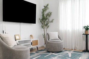 A living room with a flat-screen TV mounted on the wall, a grey armchair, a white console table, and a large potted plant.