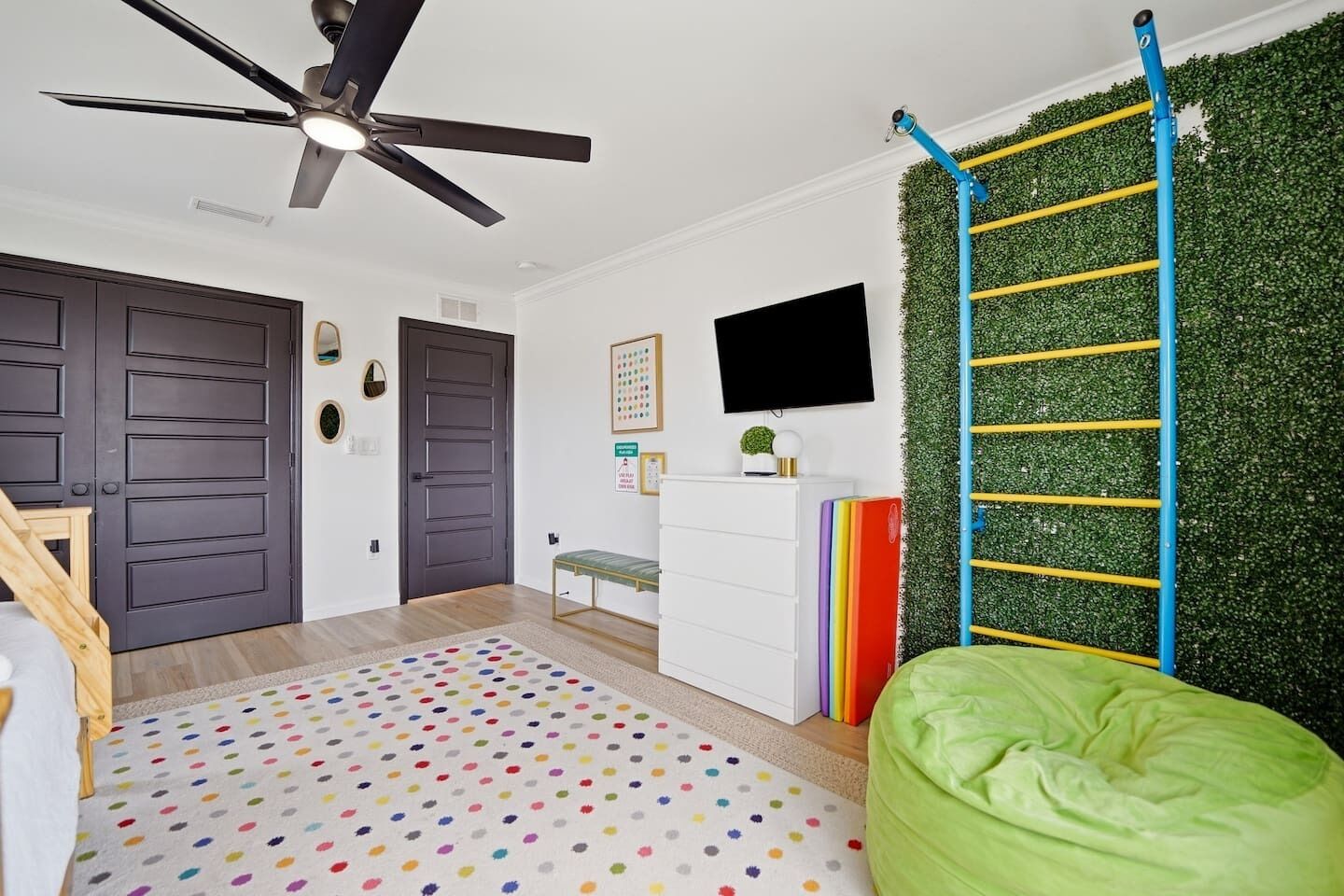 Une pièce avec un ventilateur de plafond, un mur vert agrémenté d’un mur d’escalade, un lit, un bureau et un téléviseur.